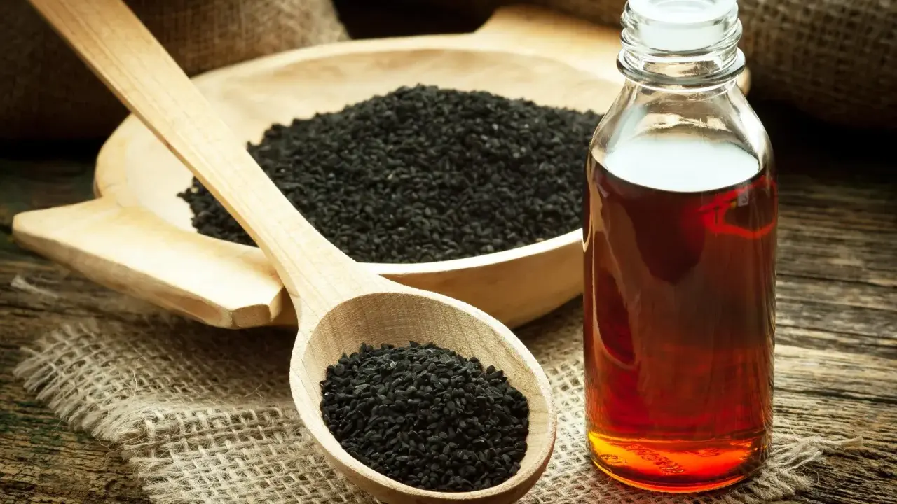 Huile de nigelle : découvrez les bienfaits pour la peau. Des graines noires dans une cuillère en bois et une bouteille d'huile ambrée sur fond rustique.