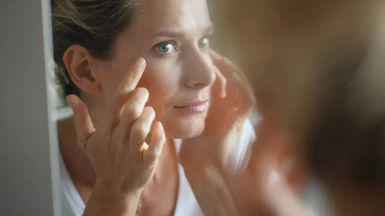 Une femme examine son visage dans un miroir, préoccupée par les signes de glycation, un processus qui affecte la peau.