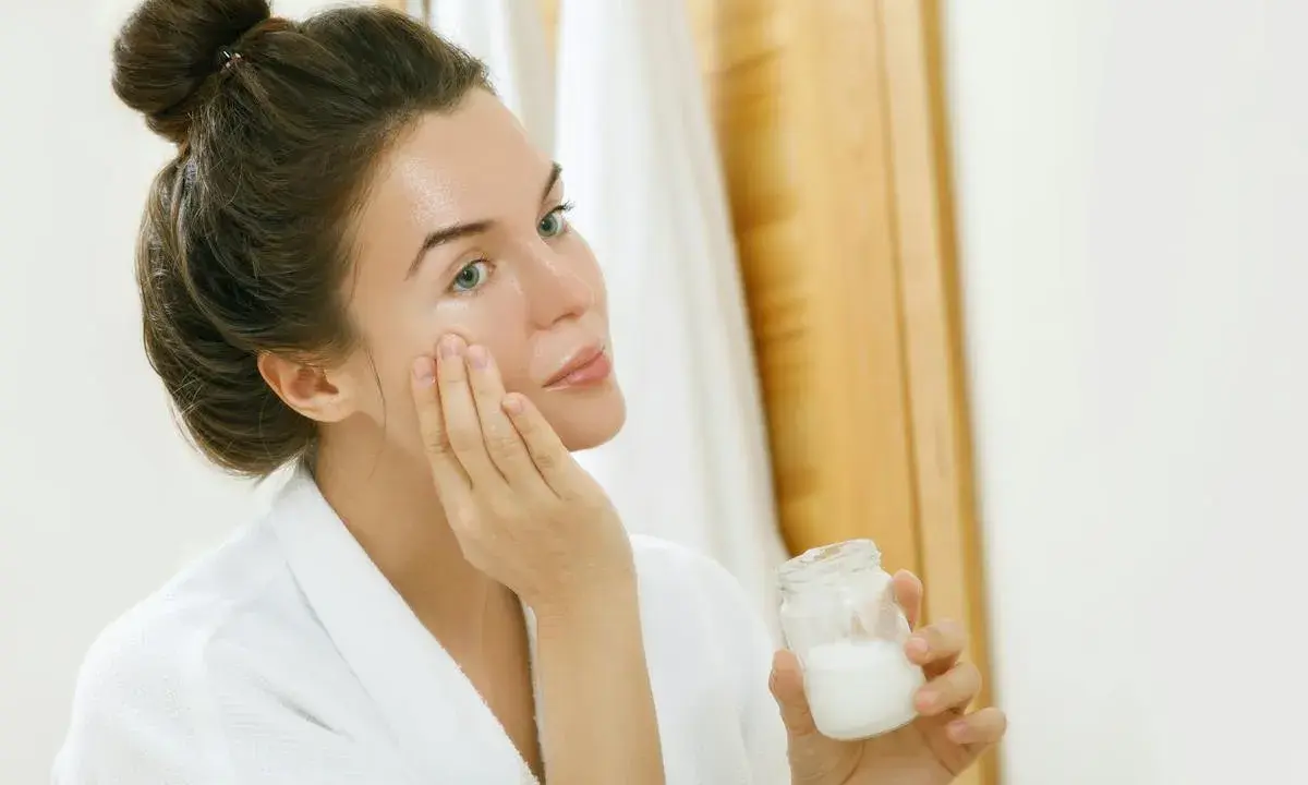 Femme appliquant une crème sur son visage, tenant un pot de ce qui pourrait être de l'huile de coco comédogène.