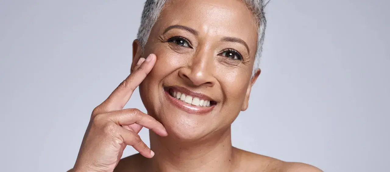 Portrait d'une femme souriante, sa peau qui marque les plis témoigne d'une vie riche. Ses doigts effleurent délicatement sa joue.
