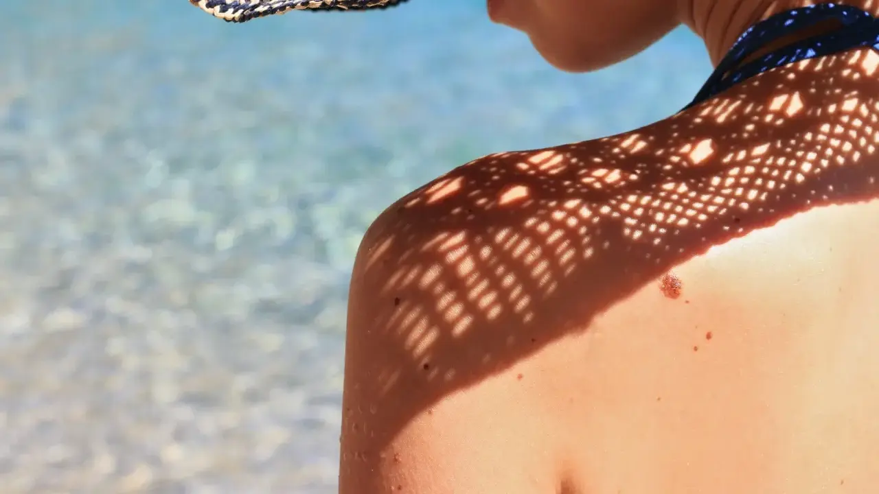 Ombre d'un chapeau sur une épaule bronzée, avec un grain de beauté soleil bien visible. L'eau bleue et claire de la mer scintille en arrière-plan.