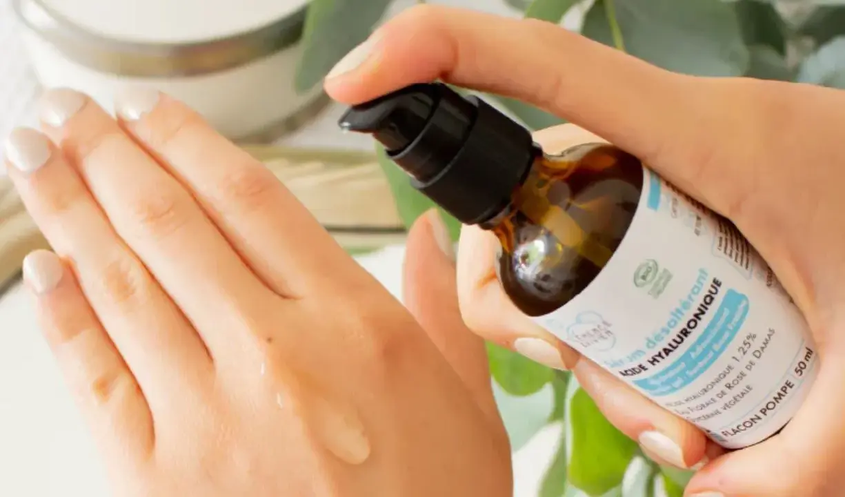 Application d'un sérum désaltérant à l'acide hyaluronique d'origine végétale sur la main. Le flacon pompe est tenu par une main aux ongles soignés.
