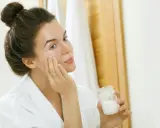 Femme appliquant une crème sur son visage, tenant un pot de ce qui pourrait être de l'huile de coco comédogène.