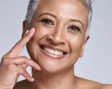 Portrait d'une femme souriante, sa peau qui marque les plis témoigne d'une vie riche. Ses doigts effleurent délicatement sa joue.