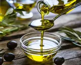 L'huile d'olive visage, dorée et pure, coule d'une cuillère dans un bol en verre. Des olives noires et des feuilles d'olivier complètent cette scène naturelle.