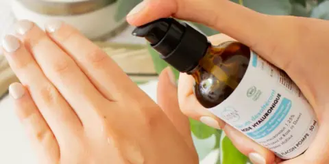 Application d'un sérum désaltérant à l'acide hyaluronique d'origine végétale sur la main. Le flacon pompe est tenu par une main aux ongles soignés.