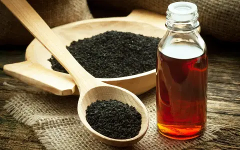 Huile de nigelle : découvrez les bienfaits pour la peau. Des graines noires dans une cuillère en bois et une bouteille d'huile ambrée sur fond rustique.