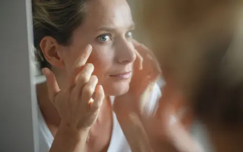 Une femme examine son visage dans un miroir, préoccupée par les signes de glycation, un processus qui affecte la peau.