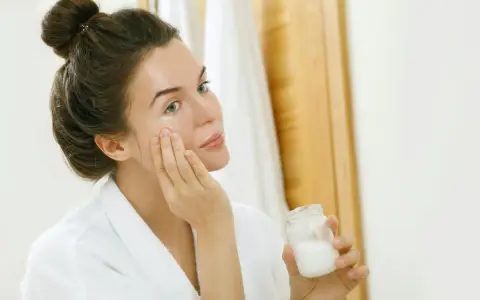 Femme appliquant une crème sur son visage, tenant un pot de ce qui pourrait être de l'huile de coco comédogène.