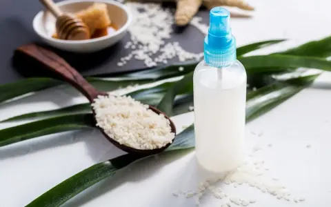Bouteille de spray contenant de l'eau de riz pour le visage, avec une cuillère de riz et du miel en arrière-plan.