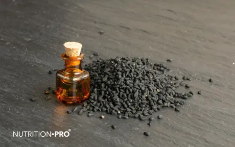 Huile de nigelle dans un flacon, à côté d'une pile de graines noires. Découvrez comment utiliser l'huile de nigelle pour ses bienfaits.