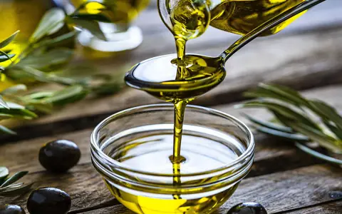 L'huile d'olive visage, dorée et pure, coule d'une cuillère dans un bol en verre. Des olives noires et des feuilles d'olivier complètent cette scène naturelle.