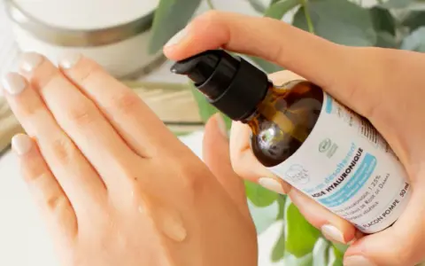 Application d'un sérum désaltérant à l'acide hyaluronique d'origine végétale sur la main. Le flacon pompe est tenu par une main aux ongles soignés.