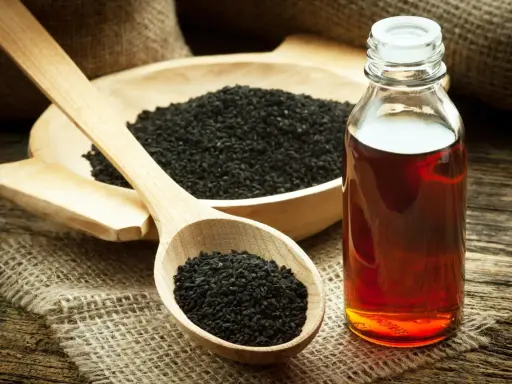 Huile de nigelle : découvrez les bienfaits pour la peau. Des graines noires dans une cuillère en bois et une bouteille d'huile ambrée sur fond rustique.