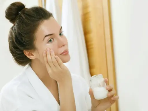 Femme appliquant une crème sur son visage, tenant un pot de ce qui pourrait être de l'huile de coco comédogène.