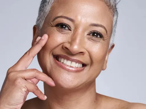 Portrait d'une femme souriante, sa peau qui marque les plis témoigne d'une vie riche. Ses doigts effleurent délicatement sa joue.