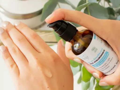 Application d'un sérum désaltérant à l'acide hyaluronique d'origine végétale sur la main. Le flacon pompe est tenu par une main aux ongles soignés.