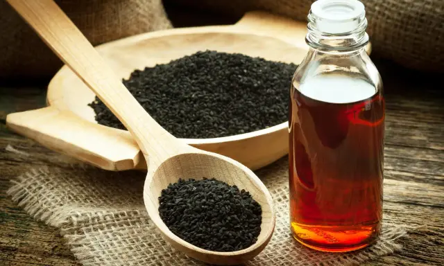 Huile de nigelle : découvrez les bienfaits pour la peau. Des graines noires dans une cuillère en bois et une bouteille d'huile ambrée sur fond rustique.