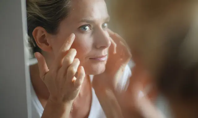 Une femme examine son visage dans un miroir, préoccupée par les signes de glycation, un processus qui affecte la peau.