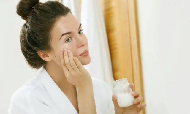 Femme appliquant une crème sur son visage, tenant un pot de ce qui pourrait être de l'huile de coco comédogène.