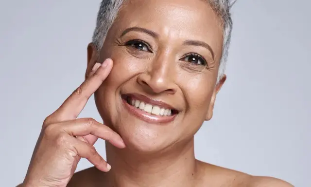 Portrait d'une femme souriante, sa peau qui marque les plis témoigne d'une vie riche. Ses doigts effleurent délicatement sa joue.