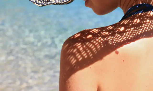 Ombre d'un chapeau sur une épaule bronzée, avec un grain de beauté soleil bien visible. L'eau bleue et claire de la mer scintille en arrière-plan.