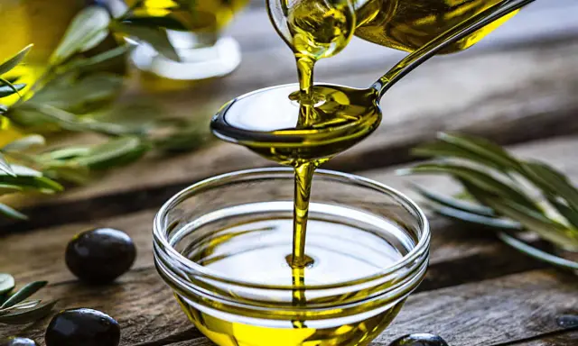 L'huile d'olive visage, dorée et pure, coule d'une cuillère dans un bol en verre. Des olives noires et des feuilles d'olivier complètent cette scène naturelle.