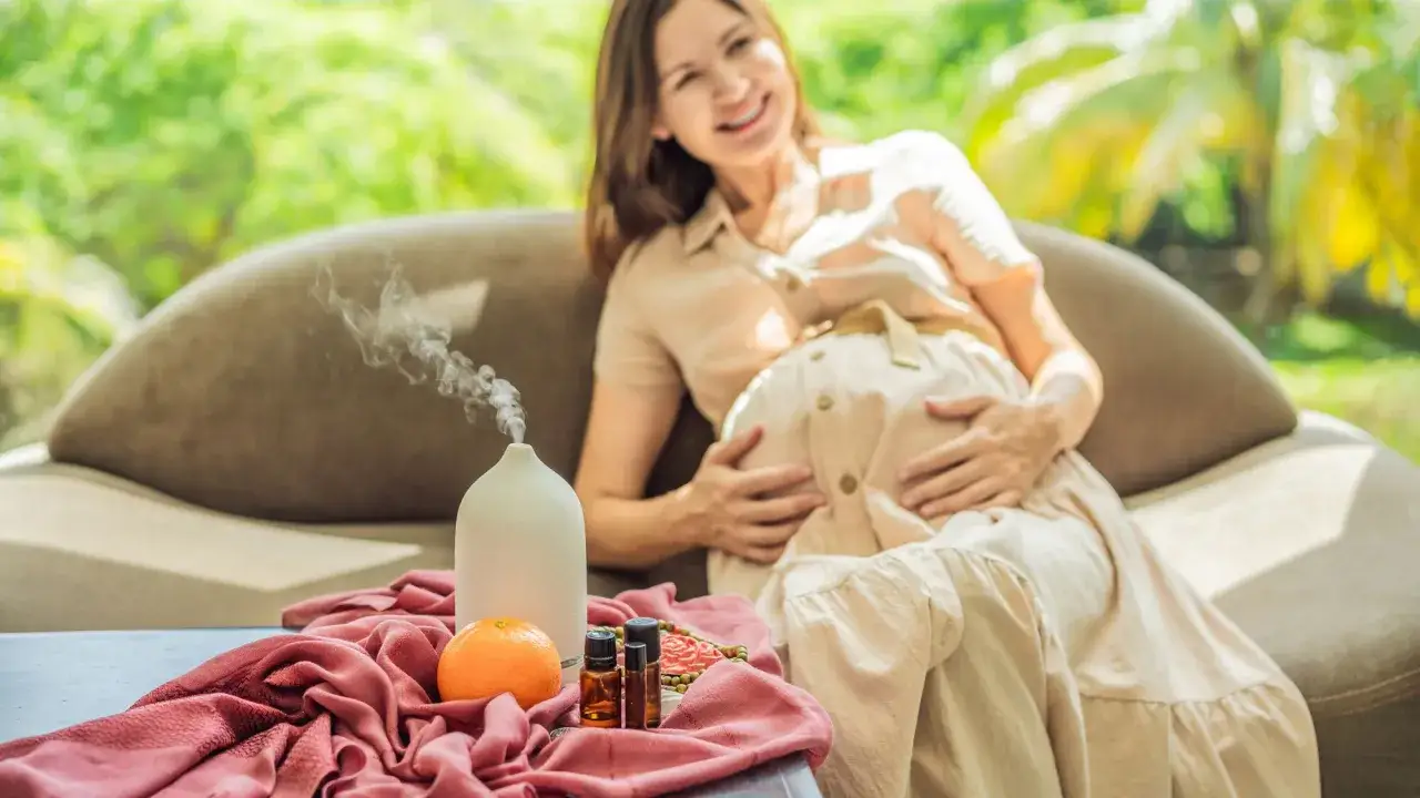 Une femme enceinte souriante se détend sur un canapé, avec un diffuseur d'huiles essentielles et une orange à proximité. L'aromathérapie peut être un allié pour une femme enceinte.