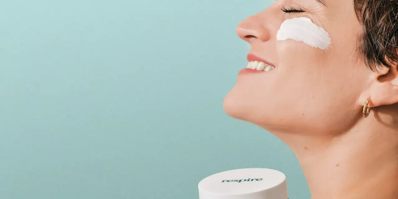 Une femme au profil souriant, appliquant une crème sur sa peau à imperfections. Le pot 
