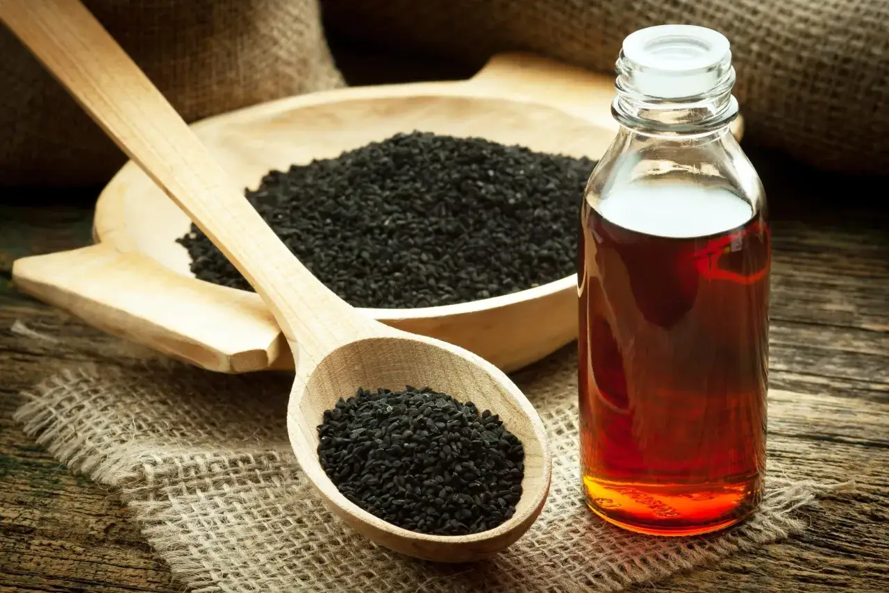 Huile de nigelle ambr&eacute;e dans un flacon, &agrave; c&ocirc;t&eacute; de graines noires dans une cuill&egrave;re en bois. D&eacute;couvrez comment utiliser l'huile de nigelle pour votre bien-&ecirc;tre.