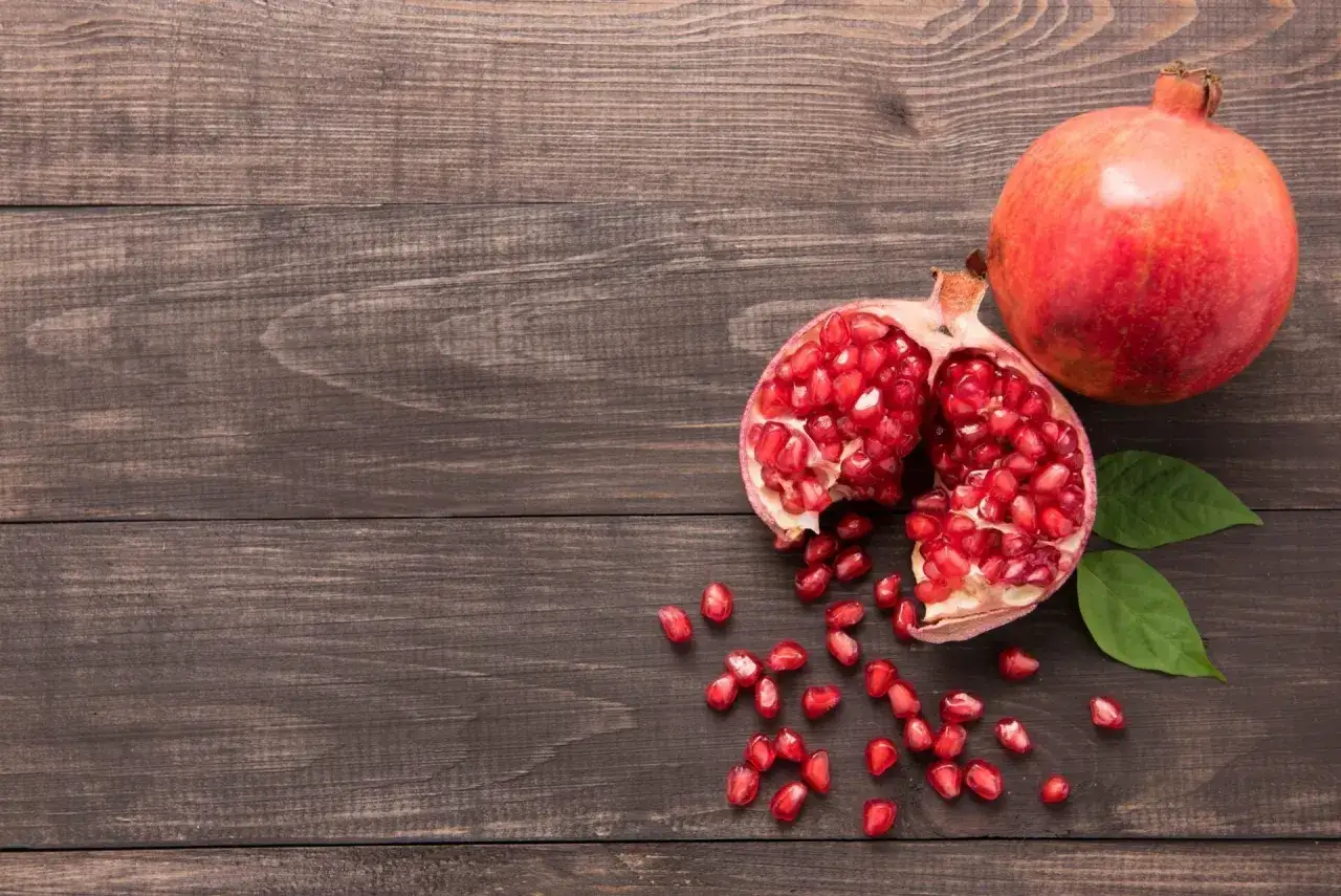 Grenade entière et ouverte, révélant ses arilles rouges. Un fruit aux multiples bienfaits pour la peau, posé sur un fond de bois sombre.